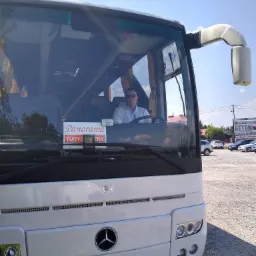 Biały autokar marki Mercedes z kierowcą w okularach za kierownicą, na przedniej szybie naklejka 'Panorama Turystyczny', w tle parking i reklama betonowych ogrodzeń.