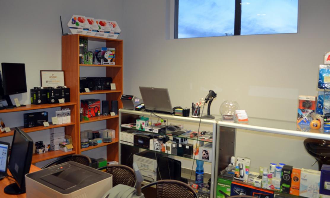 Wnętrze sklepu komputerowego z półkami pełnymi sprzętu, laptop na ladzie, drukarka na biurku i akcesoria na sprzedaż.