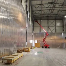 Wnętrze hali przemysłowej z metalową ścianą w trakcie montażu, widoczny podnośnik koszowy Gerken i skrzynie z materiałami.