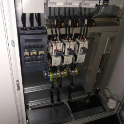 Otwarta szafa sterownicza z widocznymi stycznikami, bezpiecznikami i okablowaniem, zamontowanymi na szynach DIN.