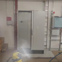 Otwarta szafa sterownicza z widocznymi elementami elektrycznymi wewnątrz, obok żółty odkurzacz przemysłowy, instalacje kablowe na ścianie, drabina serwisowa.