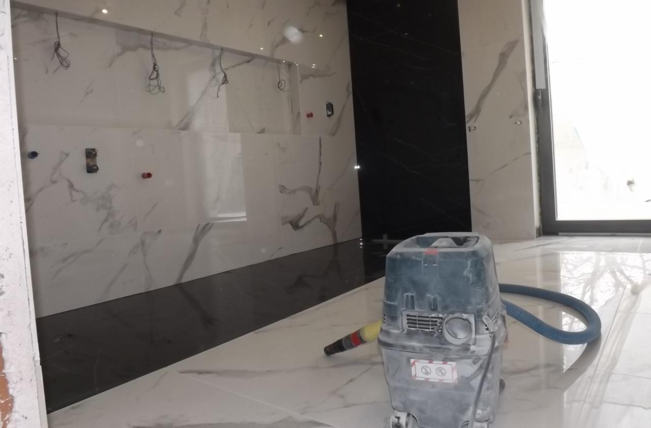 Układanie płytek marmuropodobnych z czarnym pasem, widoczny odkurzacz przemysłowy podczas sprzątania po pracach glazurniczych.