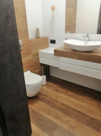 Nowoczesna łazienka z drewnopodobnymi płytkami podłogowymi i ściennymi, białą ceramiką sanitarną oraz umywalką nablatową na drewnianym blacie.