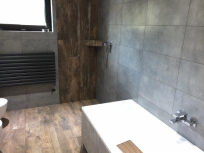 Nowoczesna łazienka z szarymi płytkami wielkoformatowymi na ścianach, drewnopodobną podłogą i elementami w kolorze drewna, grzejnik drabinkowy w kolorze grafitowym, biała wanna z baterią.