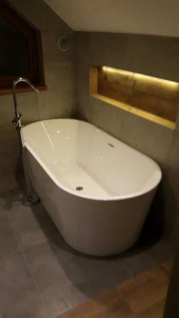 Nowoczesna łazienka z wolnostojącą białą wanną, szare płytki podłogowe i ścienne, wnęka z oświetleniem LED, bateria wannowa chromowana, okno dachowe widoczne w górnym lewym rogu.