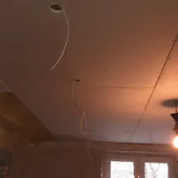 Montaż płyt kartonowo-gipsowych na suficie, widoczne przewody elektryczne wychodzące przez otwory, okno z widokiem na zimowy krajobraz.