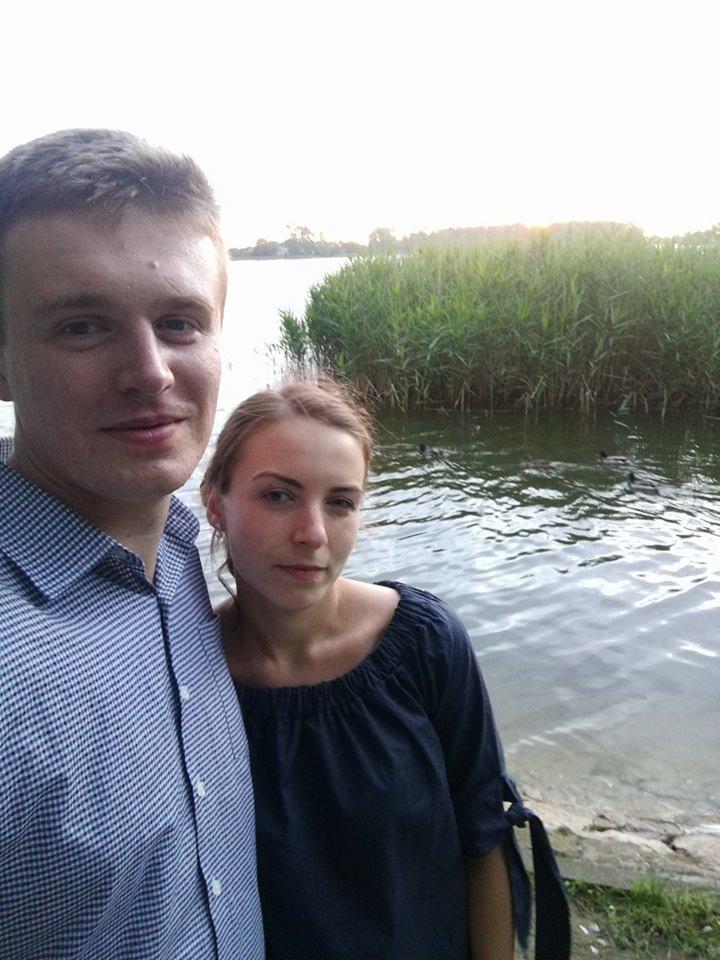 Para pozuje na tle jeziora z trzcinami o zmierzchu. Mężczyzna w koszuli w kratę, kobieta w granatowej bluzce. Lekki uśmiech, naturalne światło.