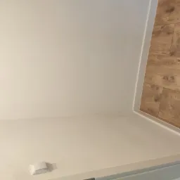 Świeżo pomalowane jasne ściany z widocznym włącznikiem elektrycznym i fragmentem drewnopodobnej podłogi ułożonej w jodełkę.