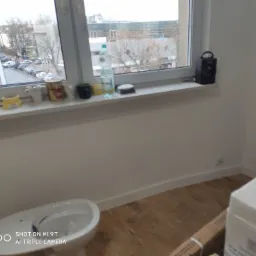 Biała miska ustępowa na drewnianej podłodze w trakcie instalacji, widoczne pudełka i akcesoria na parapecie okna z widokiem na ulicę.