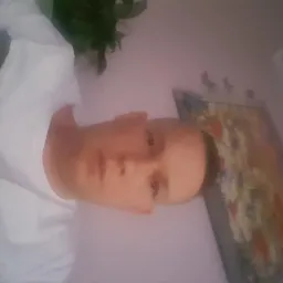 Portret mężczyzny w białej koszulce na tle ściany z obrazem i rośliną doniczkową, obrócony o 90 stopni w lewo.