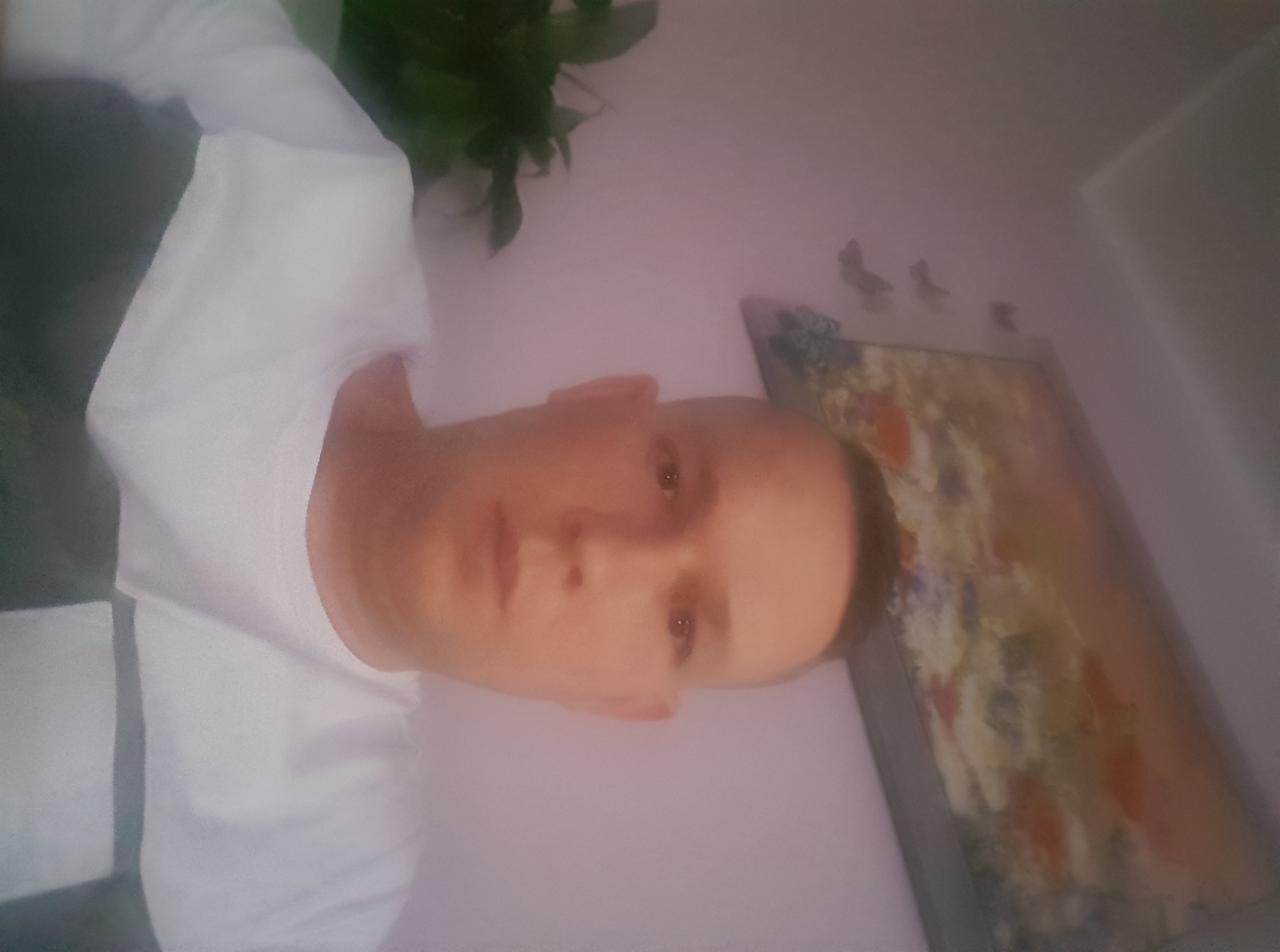 Portret mężczyzny w białej koszulce na tle ściany z obrazem i rośliną doniczkową, obrócony o 90 stopni w lewo.