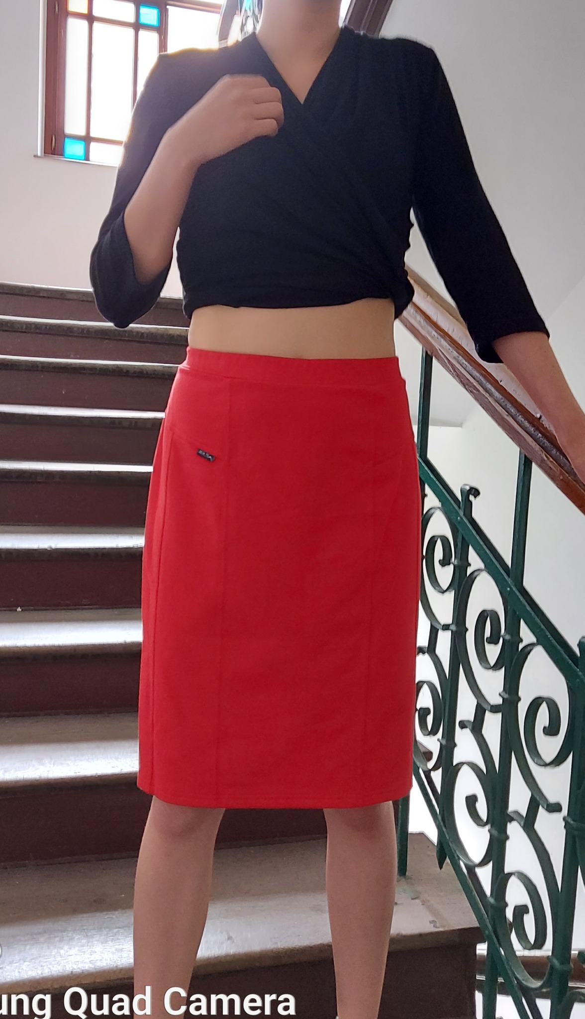 Kobieta prezentuje czerwoną spódnicę ołówkową z małą kieszonką na schodach, ubrana w czarną bluzkę kopertową.