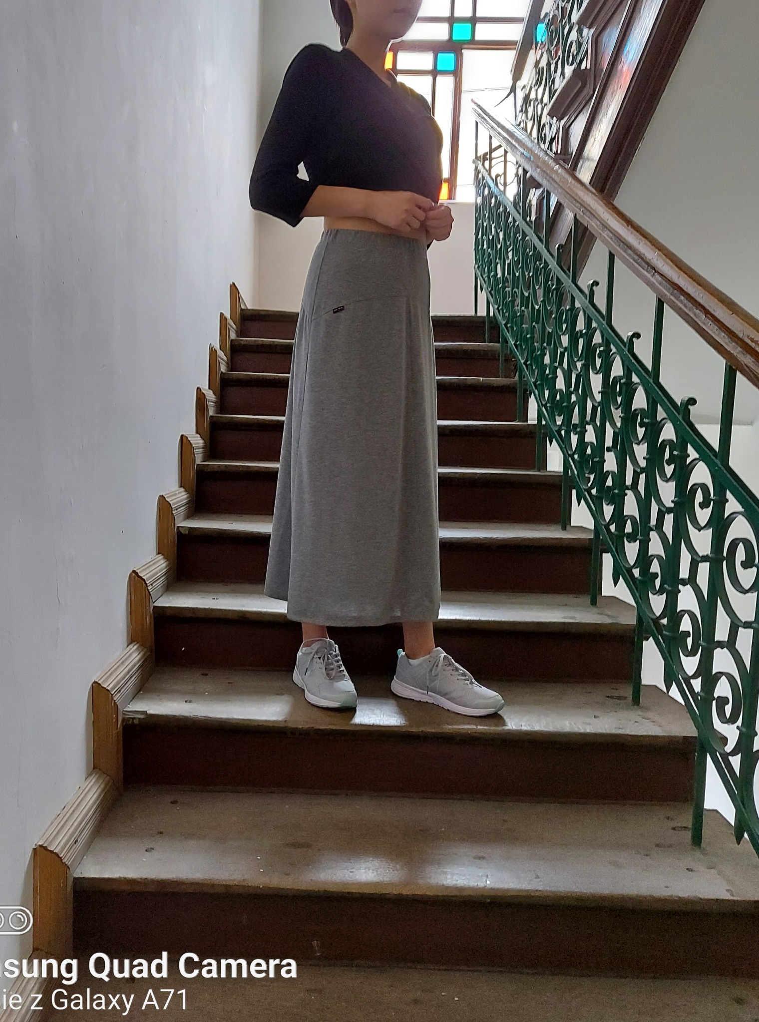 Kobieta pozuje na schodach w szarej, długiej spódnicy dresowej i czarnej bluzce z długim rękawem, widoczne szare sportowe buty i zdobiona, zielona balustrada schodów.