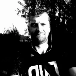 Czarno-białe selfie mężczyzny z słuchawkami na tle drzew i domu. Oświetlenie kontrastowe, widoczna twarz i fragment koszulki.