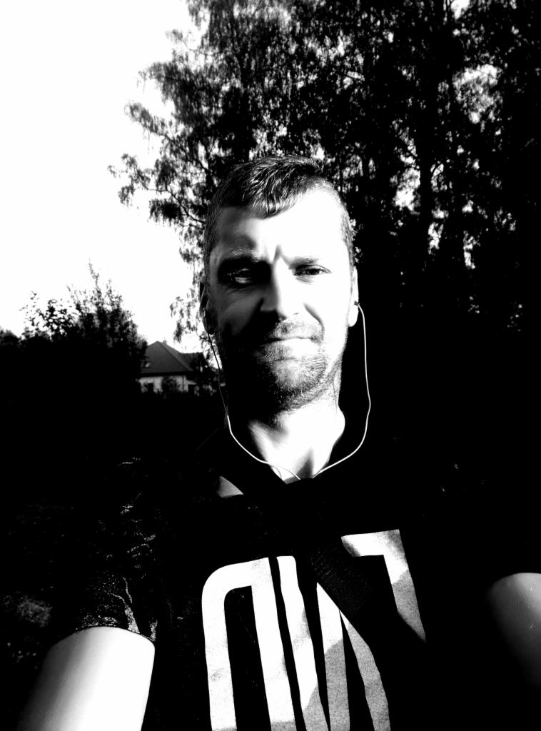 Czarno-białe selfie mężczyzny z słuchawkami na tle drzew i domu. Oświetlenie kontrastowe, widoczna twarz i fragment koszulki.