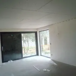 Surowe wnętrze pokoju z zamontowaną płytą kartonowo-gipsową na suficie, duże przesuwne okno z widokiem na zewnątrz, ślady po instalacji elektrycznej na ścianie.