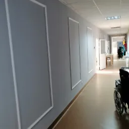 Korytarz w plac&oacute;wce medycznej z szarymi ścianami ozdobionymi białymi, prostokątnymi listwami dekoracyjnymi; na końcu korytarza widoczne otwarte drzwi i krzesło inwalidzkie.