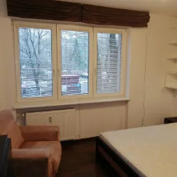 Pokój z beżową sofą, łóżkiem z ciemną ramą i oknem z żaluzjami, nad którym wisi brązowa roleta rzymska.