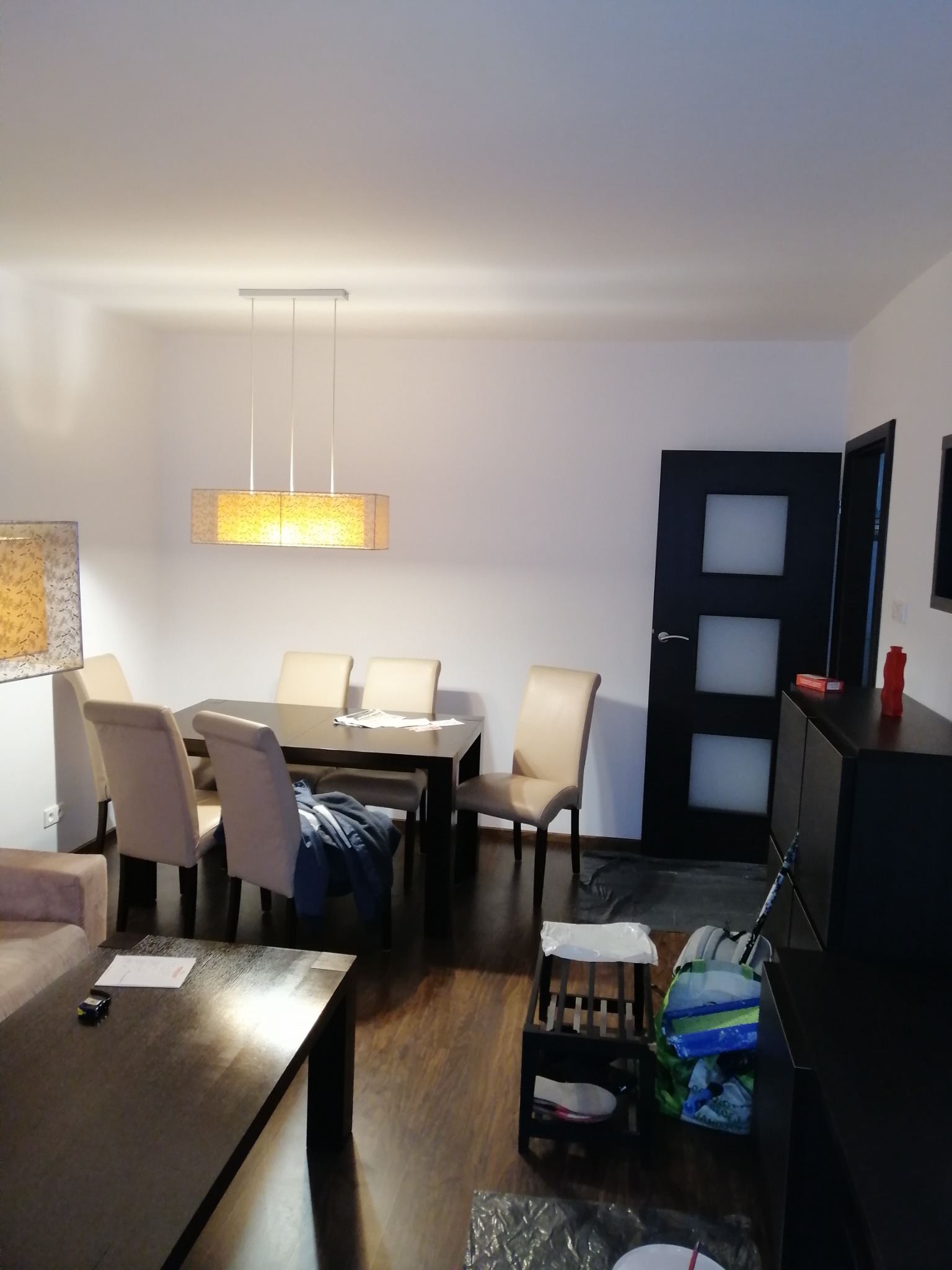 Jasne wnętrze pokoju dziennego z drewnianą podłogą, stołem jadalnym pod wiszącą lampą i czarnymi meblami w tle, w tym drzwi z białymi panelami.