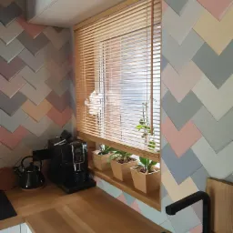 Jasne drewniane żaluzje poziome w oknie kuchennym z kolorową, wzorzystą mozaiką na ścianie, rośliny w drewnianych doniczkach na parapecie, ekspres do kawy i czajnik na blacie.