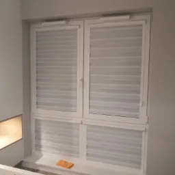 Dwuskrzydłowe okno z zamontowanymi, częściowo zasłoniętymi roletami w odcieniach bieli i szarości; na parapecie leży pomarańczowa ściereczka.