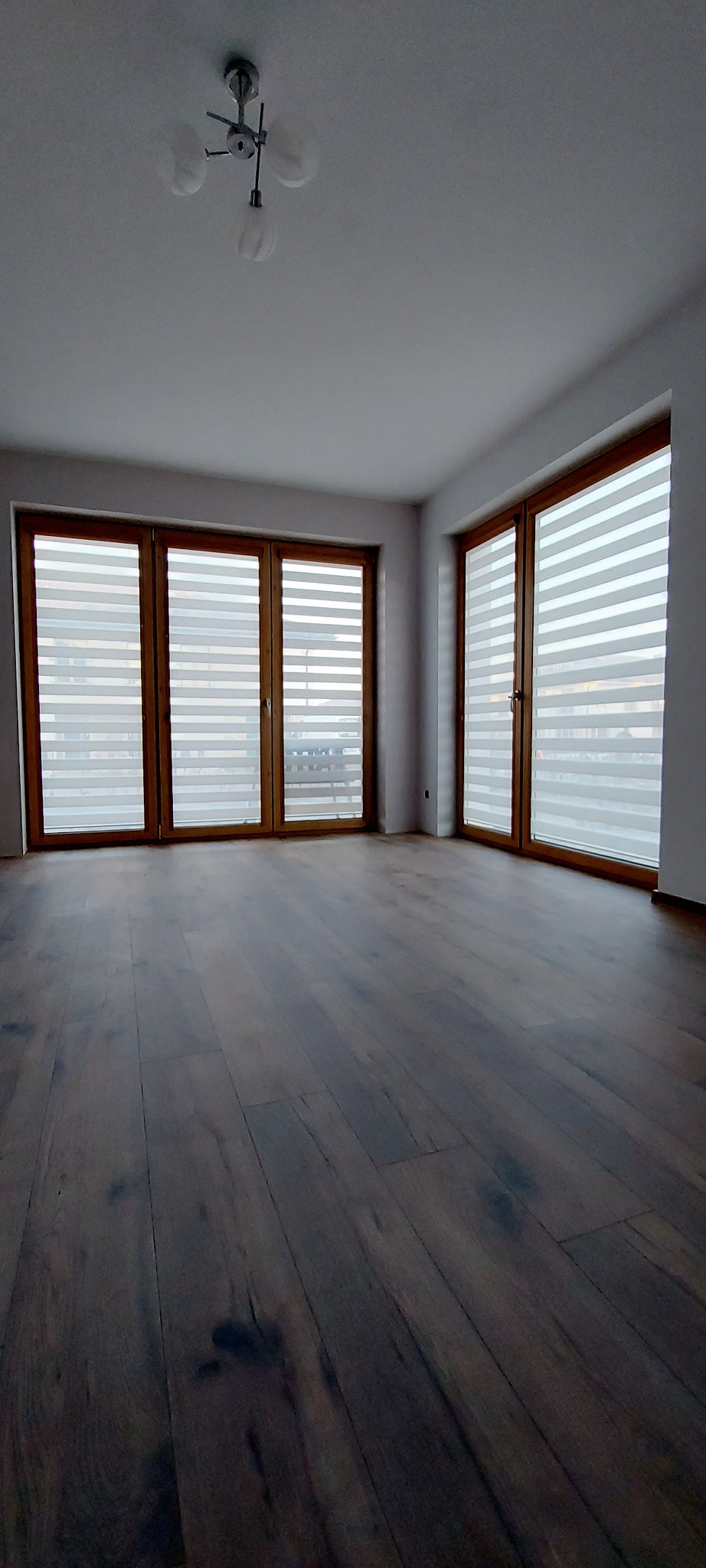 Wnętrze pokoju z trzema oknami wyposażonymi w poziome przesłony okienne z naprzemiennymi pasami transparentnymi i zaciemniającymi, widok na parkiet z ciemnego drewna.