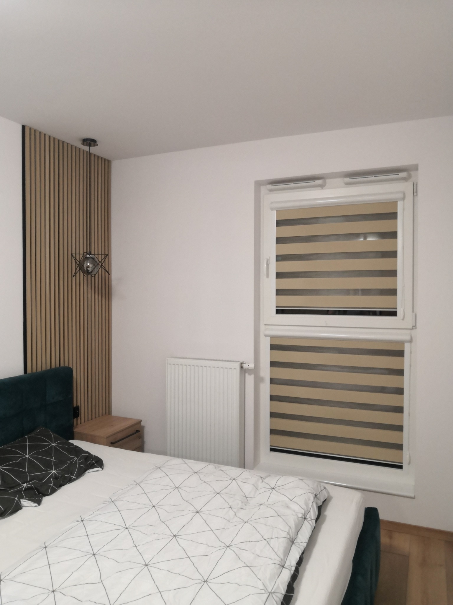 Sypialnia z beżowo-szarymi roletami w oknie, ustawionymi w poziome pasy, widoczne drewniane panele na ścianie i nowoczesna lampa wisząca.