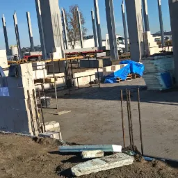 Budowa hali: widok na wznoszone ściany z betonu komórkowego, stalowe słupy konstrukcyjne i przygotowane zbrojenie. W tle widoczny sprzęt budowlany i zaparkowane ciężarówki.