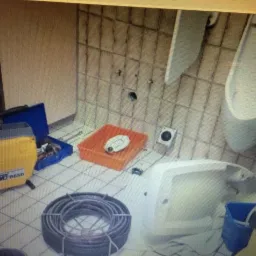 Rozmontowane pisuary w łazience z częściowo zdjętymi płytkami, obok narzędzia hydrauliczne, w tym zwinięty wąż do udrażniania.