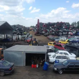 Widok na plac z wrakami samochodów, stertę pojazdów przygotowanych do recyklingu, dźwig i budynki gospodarcze w słoneczny dzień.