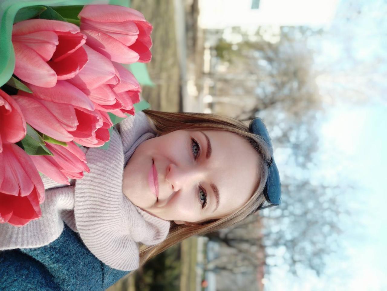 Uśmiechnięta blondynka z bukietem różowych tulipanów na tle drzew i błękitnego nieba. Kobieta ma okulary na głowie i szal na szyi. Jasne, naturalne światło.