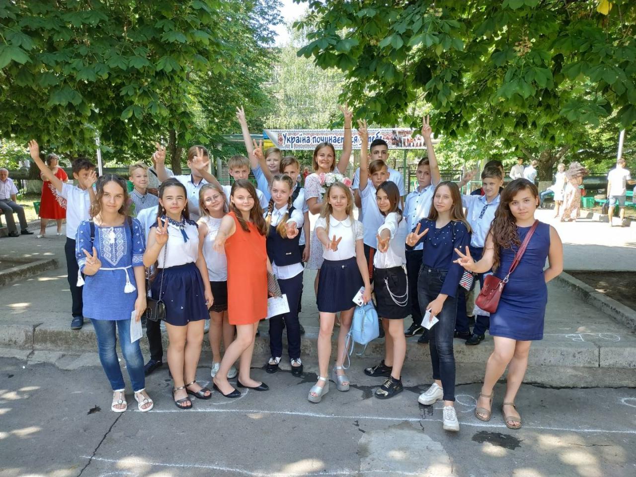 Grupa nastolatków i dorosła kobieta pozują do zdjęcia na zewnątrz, trzymając ręce w geście 'V', z napisem w tle 'Ukraina zaczyna się'.