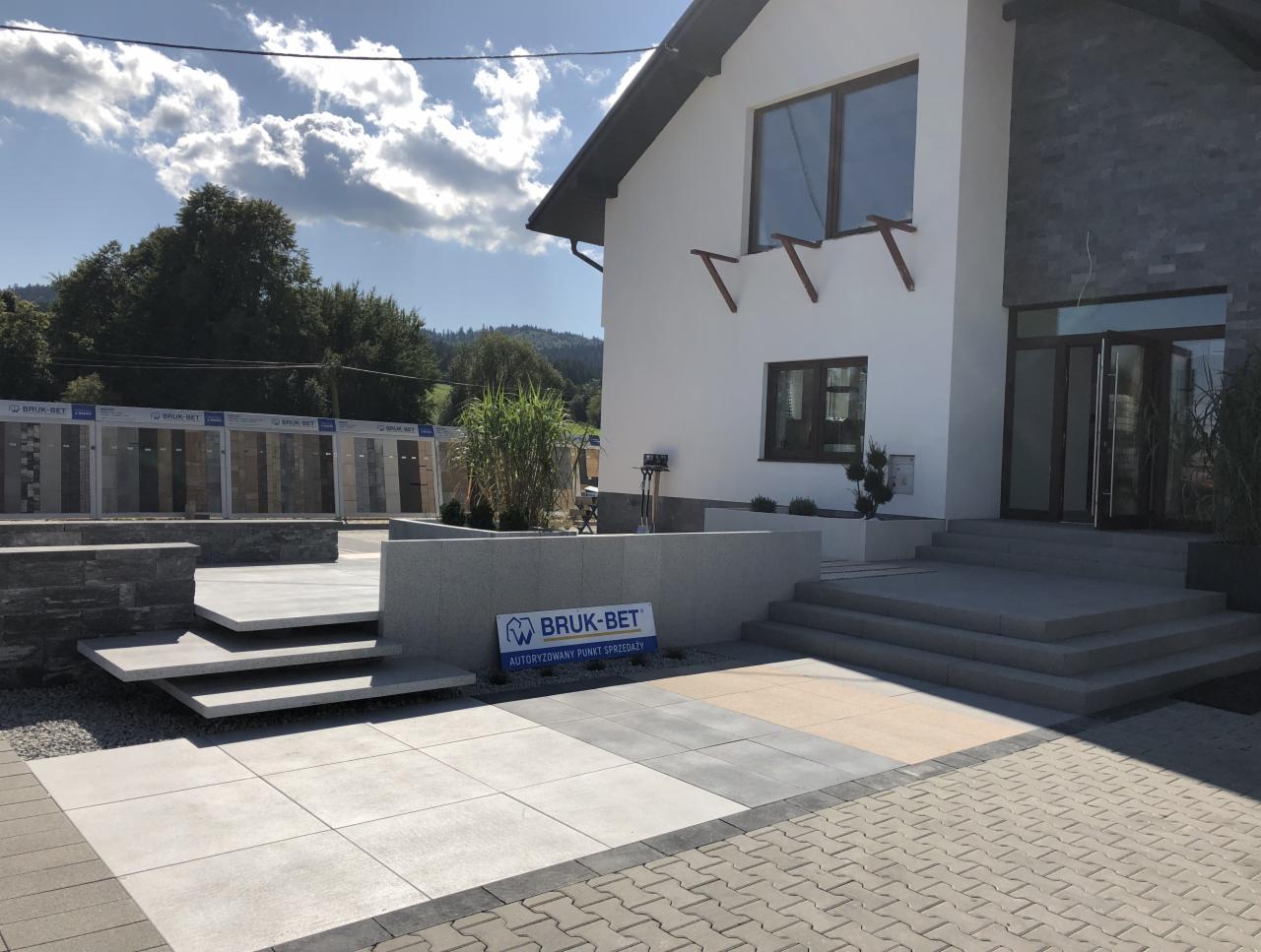 Ekspozycja różnych rodzajów nawierzchni z kostki brukowej przed budynkiem z elementami kamiennymi i logo producenta.