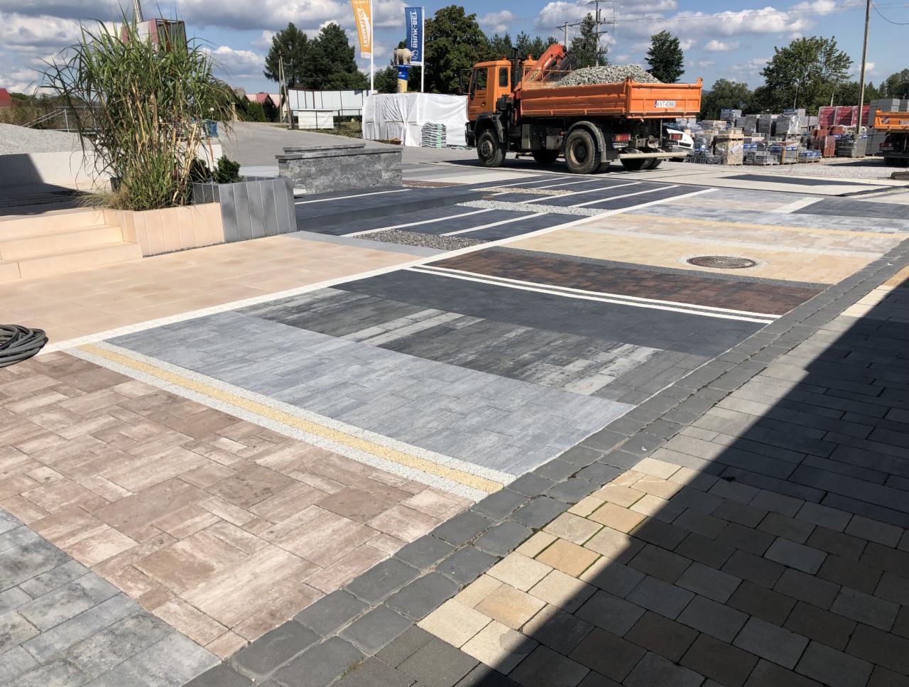 Ekspozycja różnych wzorów i kolorów nawierzchni z kostki brukowej na placu, z pomarańczową ciężarówką w tle i paletami kostki w oddali.