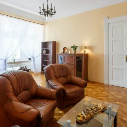 Jasne biuro z dwoma skórzanymi fotelami, biurkiem, regałem i stolikiem kawowym z przekąskami, oświetlone przez żyrandol i naturalne światło z okna.