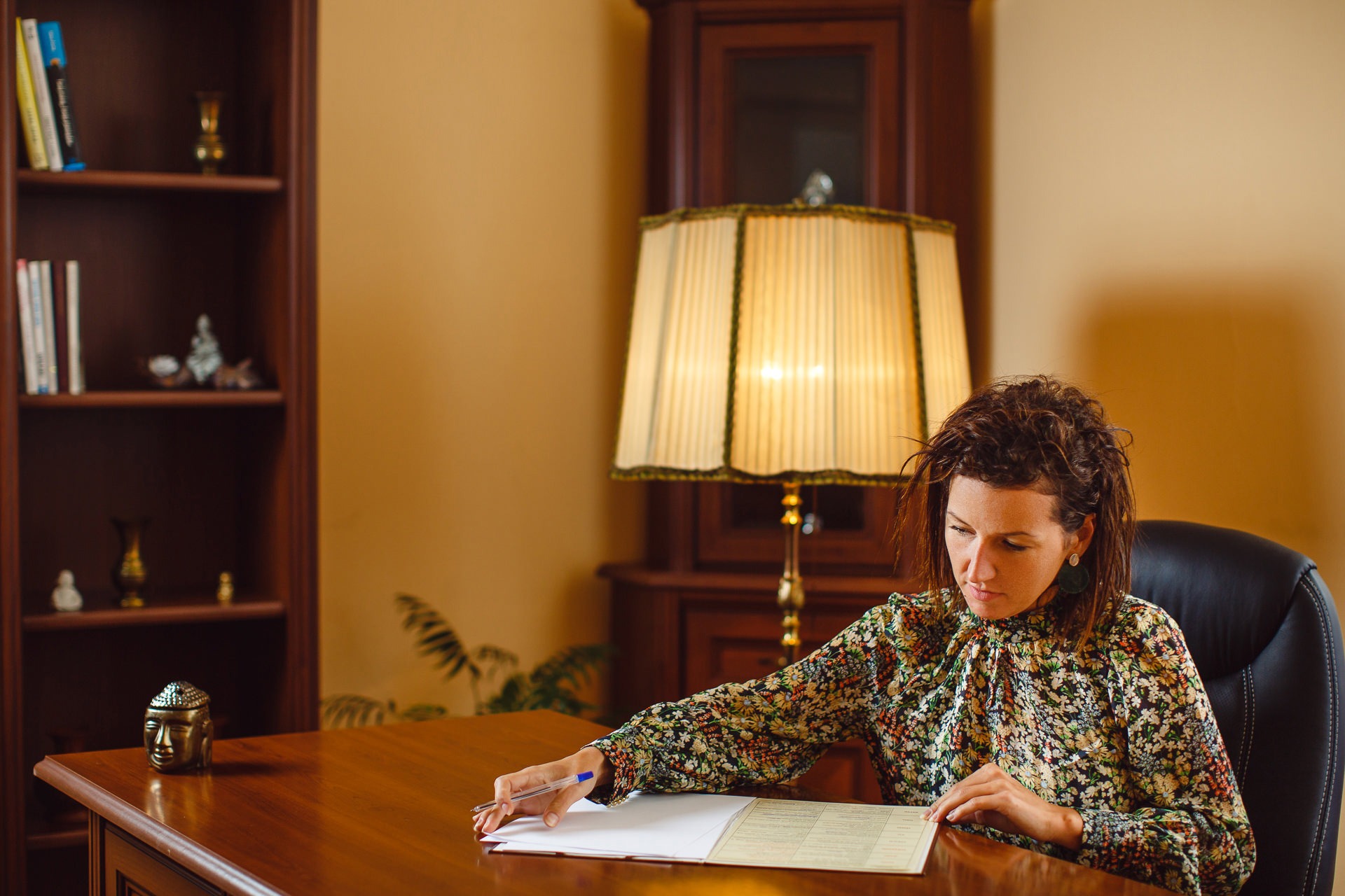 Kobieta w kwiecistej bluzce siedzi przy biurku z drewnianym blatem, wypełniając dokument. Na biurku leży długopis, a w tle widoczny regał z książkami i lampa ze złotą podstawą.