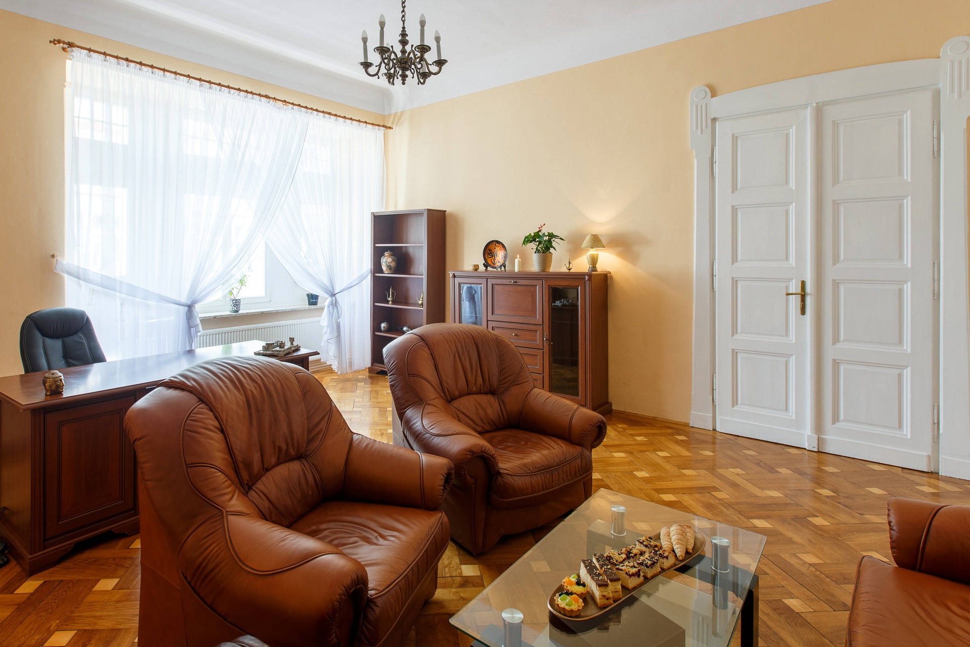 Jasne biuro z dwoma skórzanymi fotelami, biurkiem, regałem i stolikiem kawowym z przekąskami, oświetlone przez żyrandol i naturalne światło z okna.