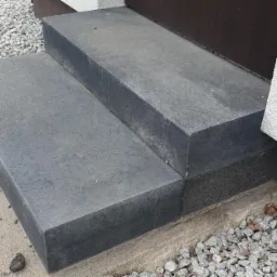 Dwa stopnie schodowe wykonane z ciemnoszarej kostki granitowej prowadzące do drzwi wejściowych, z widocznym fragmentem elewacji budynku o jasnej strukturze i podsypką z drobnego kamienia.