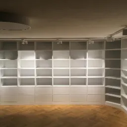 Biała, pusta biblioteka na wymiar z zabudowanymi szafkami dolnymi i oświetleniem punktowym na szynoprzewodach, parkiet w jodełkę na podłodze.