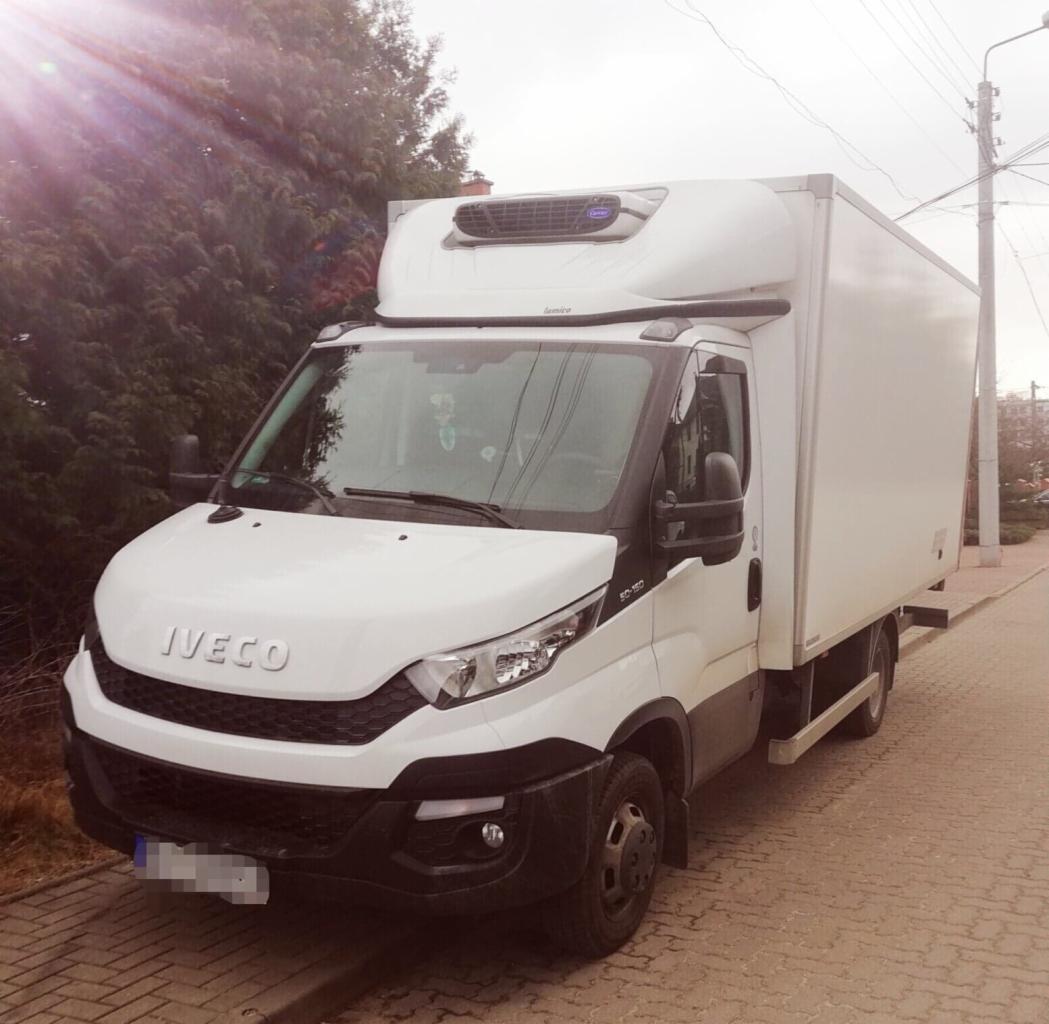 Biały samochód dostawczy Iveco z chłodnią, zaparkowany na chodniku z kostki brukowej, widok z przodu, z widocznym drzewem w tle.