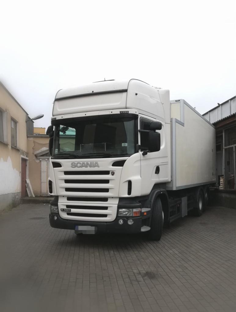 Biała ciężarówka Scania z zabudową skrzyniową zaparkowana na brukowanym placu, widoczna z przodu pod lekkim kątem, w tle budynki.