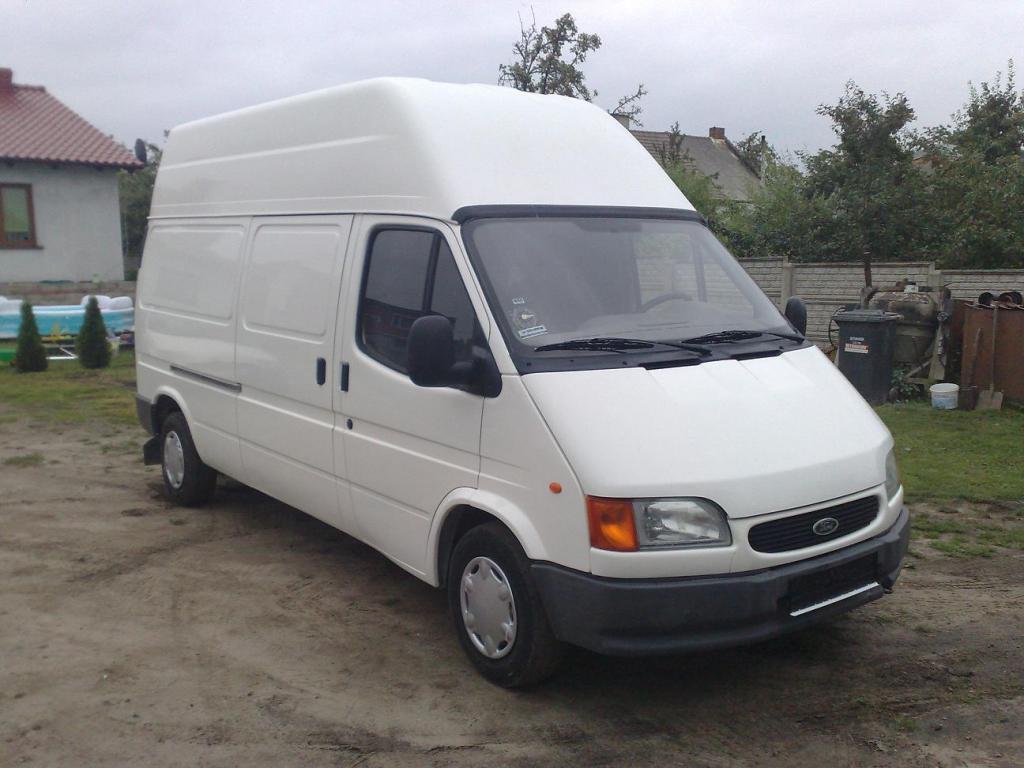 Biały, podwyższony samochód dostawczy Ford Transit na ziemistym podłożu, z widocznym fragmentem domu z czerwoną dachówką w tle.