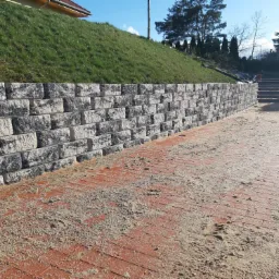 Mur oporowy z szarej kostki przytarasowej, zabezpieczający zielony nasyp, z elementami czerwonej kostki brukowej na chodniku, częściowo zasypanej piaskiem.
