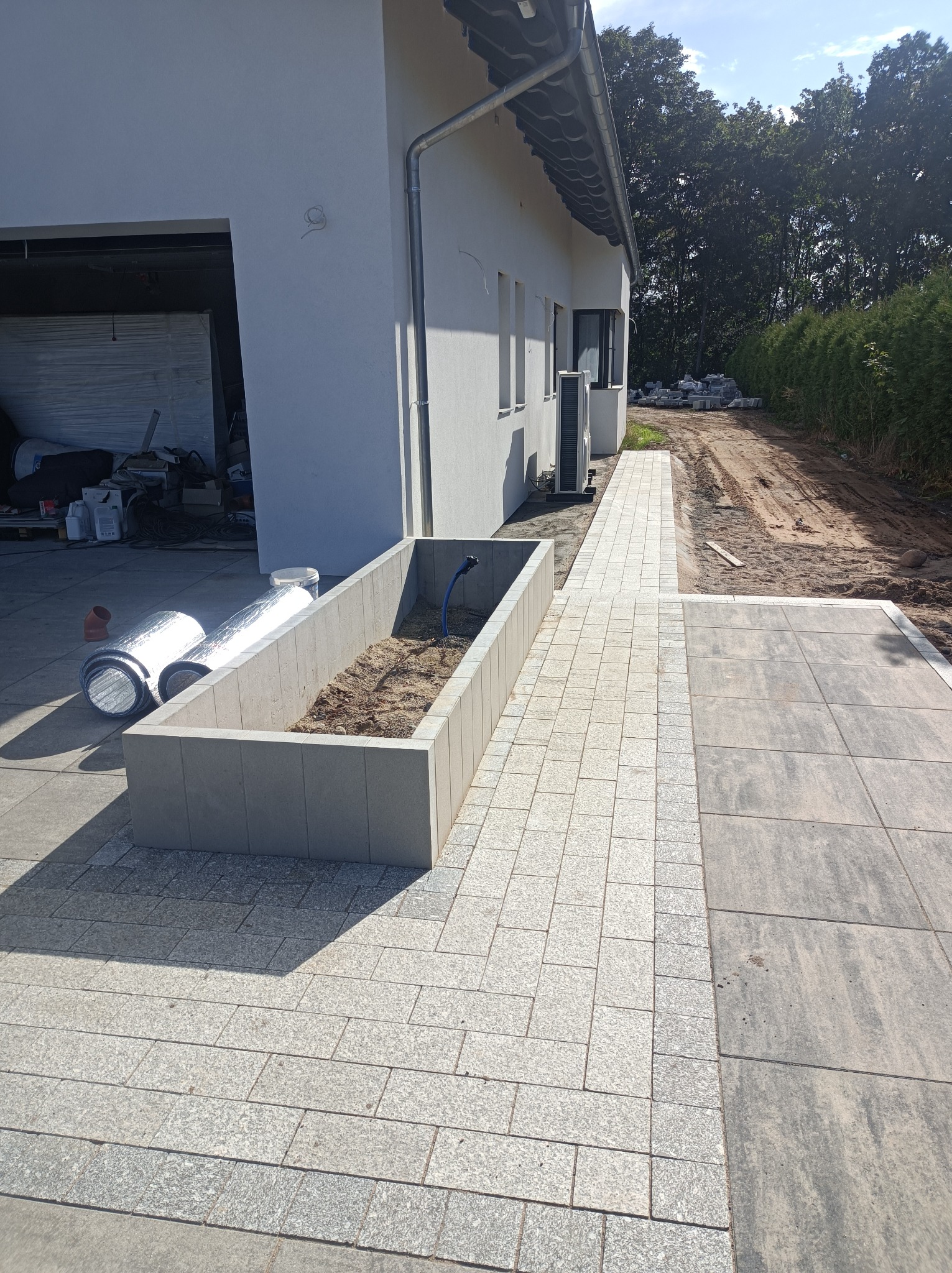 Świeżo ułożona kostka granitowa w odcieniach szarości, obok białego domu z garażem, widoczna rabata z ziemią i wężykiem do podlewania, w tle przygotowany teren pod dalsze prace.