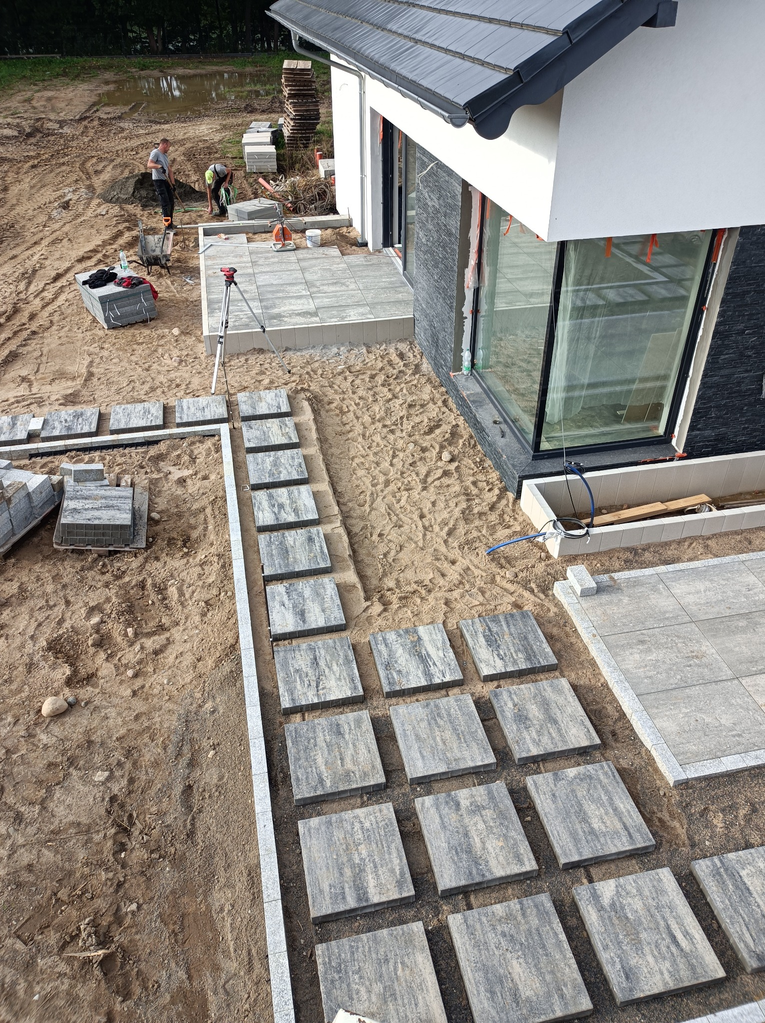 Montaż ścieżki z płyt betonowych w ogrodzie, widoczne płyty ułożone na piasku, obrzeża z kostki granitowej, w tle pracownicy przygotowujący podłoże, dom z dużymi oknami.