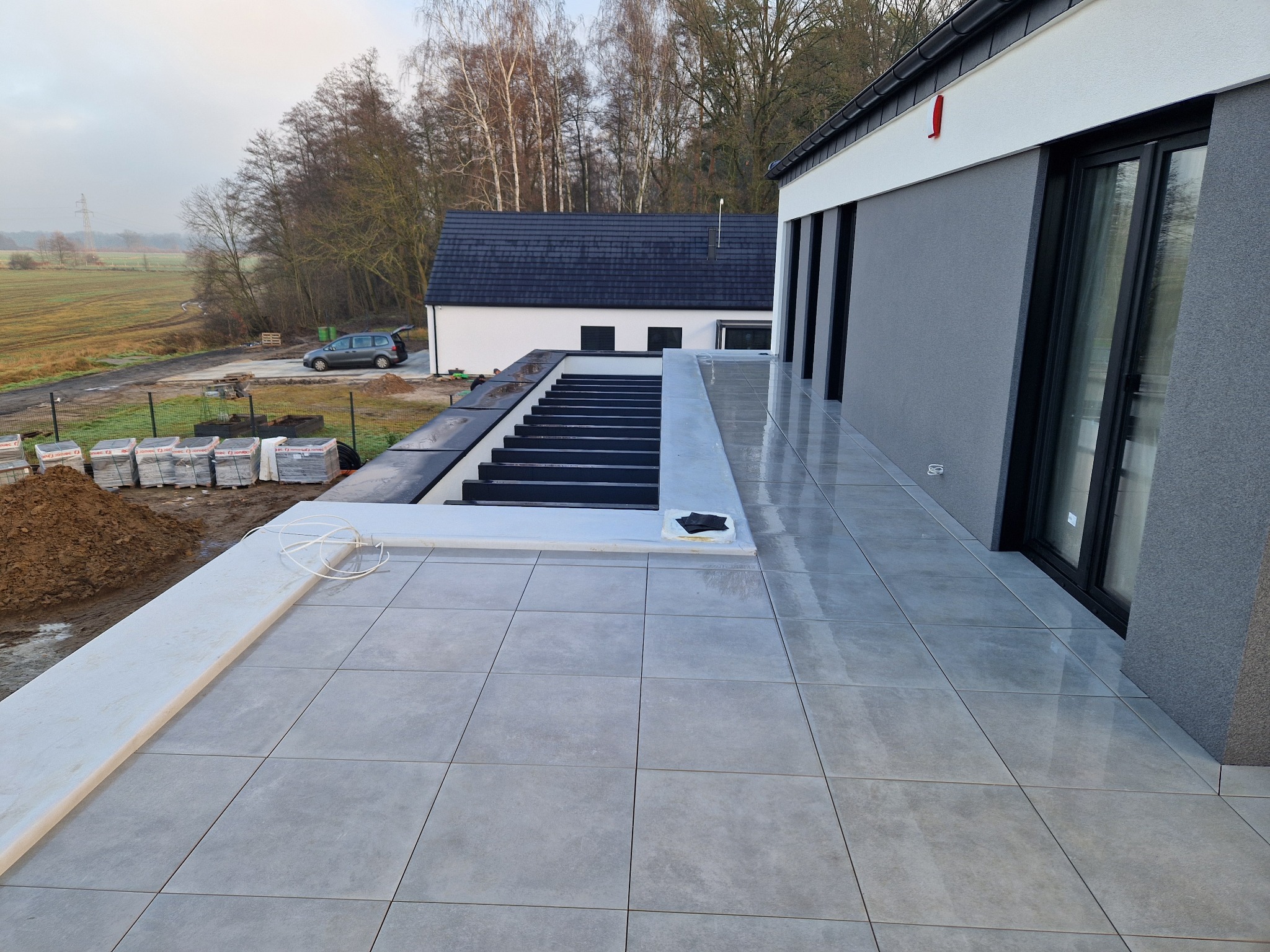 Nowoczesny taras z szarych płyt, widok z góry. W tle dom z dużymi oknami i przygotowane podłoże pod dalszą część tarasu. Na ziemi sterta kostki brukowej.