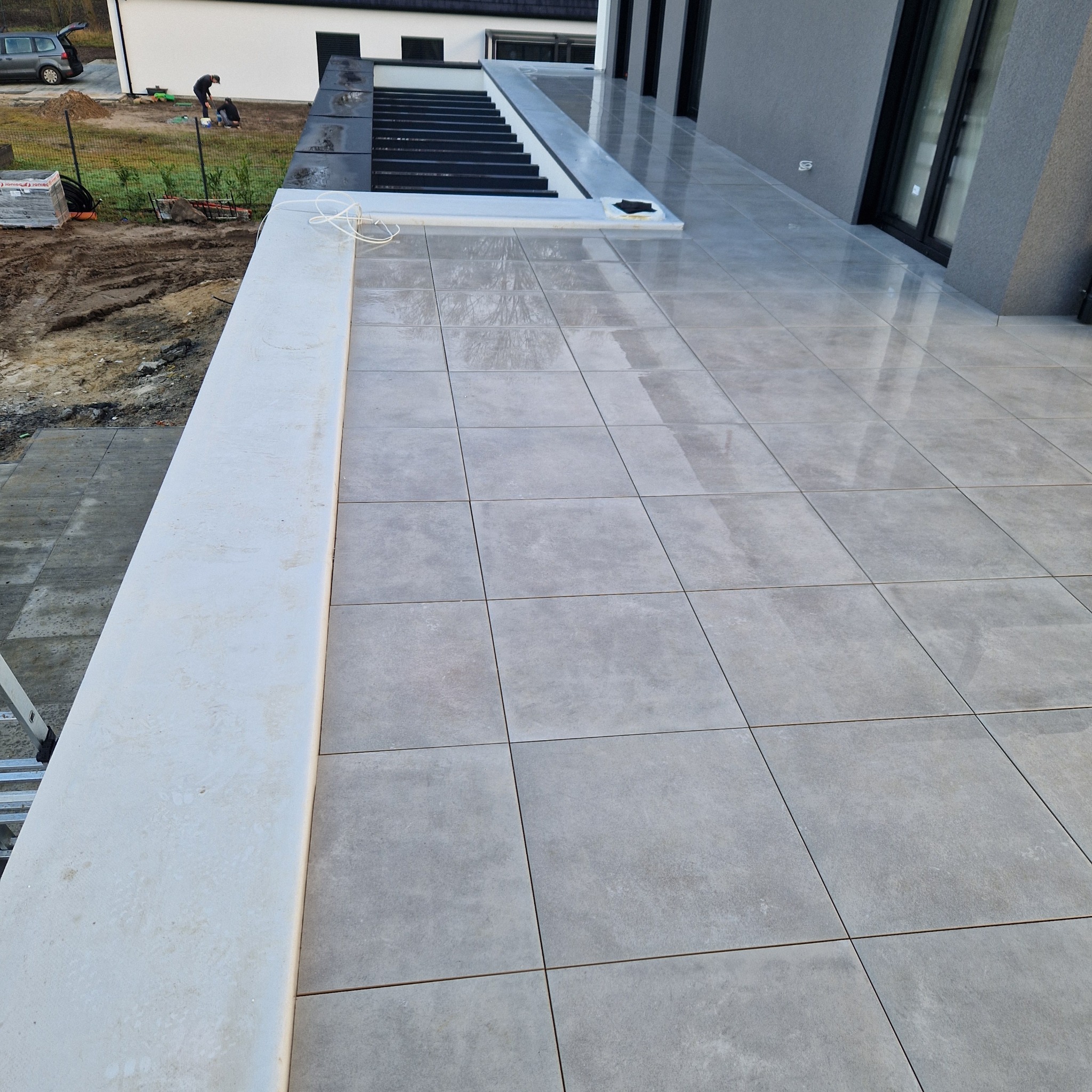 Wykończony taras z szarych, kwadratowych płyt. Widoczne detale: białe obrzeże, czarne schody i fragment budynku w tle. Prace brukarskie na zewnątrz.