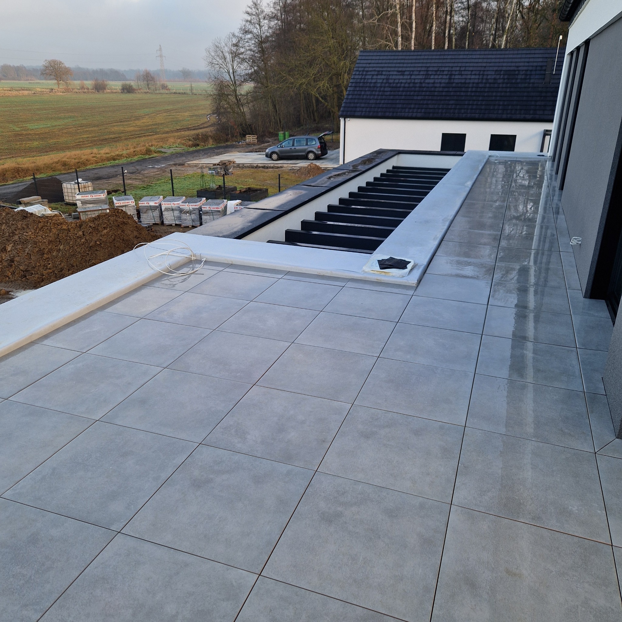 Nowoczesny taras z szarych płyt, z widokiem na okolicę. Na tarasie widoczne materiały budowlane, sugerujące prace wykończeniowe. Minimalistyczny design i świeże powietrze.