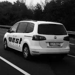 Biały van marki Volkswagen z napisem 'West Service' na boku, jadący autostradą, widok z tyłu pod kątem, czarno-białe zdjęcie.