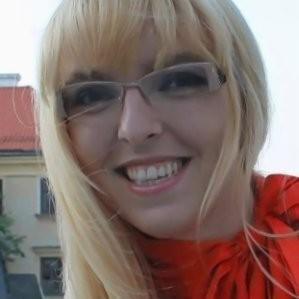 Portret uśmiechniętej kobiety w okularach, z rozwianymi blond włosami, na tle miejskiej zabudowy z elementami czerwonego dachu.